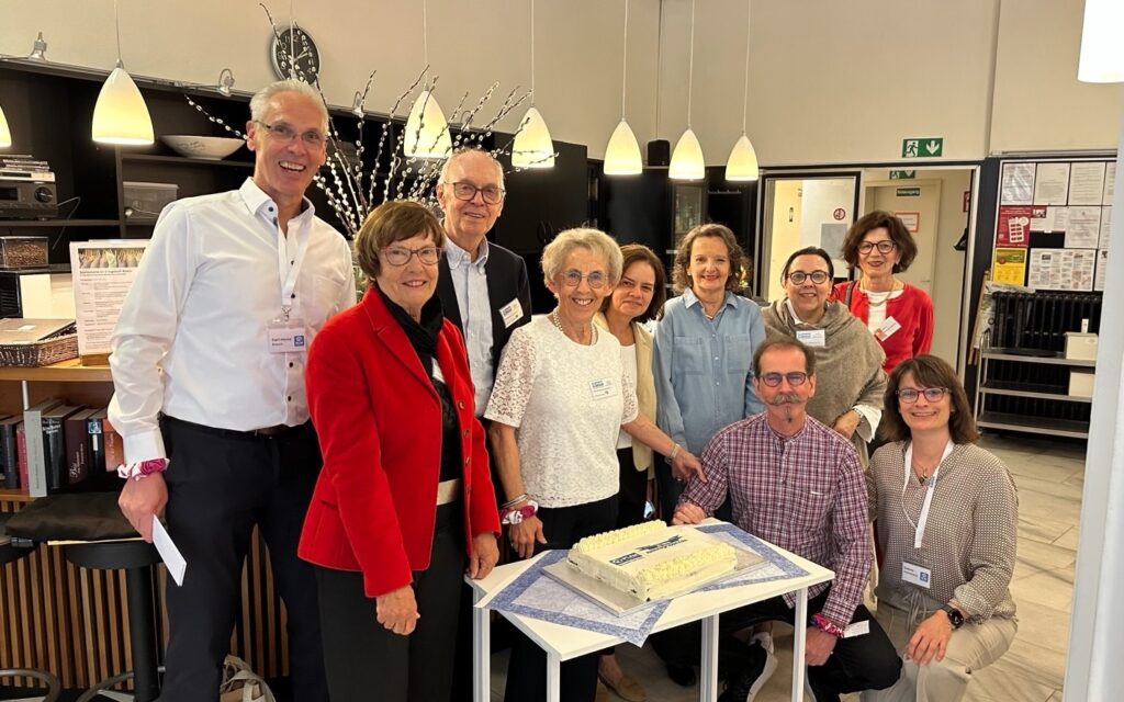 A group of ten adults, smiling and posing around a table with a rectangular cake; they are indoors under hanging lights, with a kitchen area in the background.