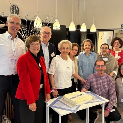 A group of ten adults, smiling and posing around a table with a rectangular cake; they are indoors under hanging lights, with a kitchen area in the background.