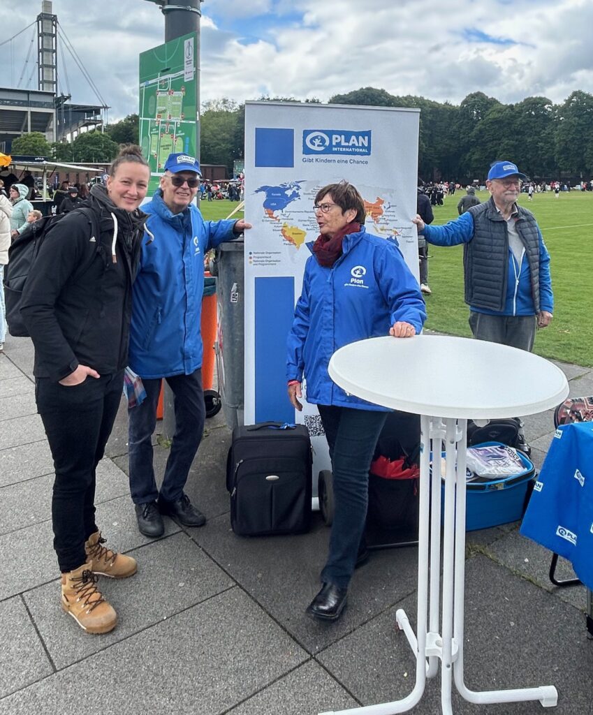 Vier Personen stehen bei einer Veranstaltung im Freien neben einem runden Tisch und einer Anzeigetafel für PLAN International, mit einem Sportplatz, einer Menschenmenge und Bäumen im Hintergrund. Zwei Personen tragen blaue Jacken und Hüte mit dem PLAN-Logo.