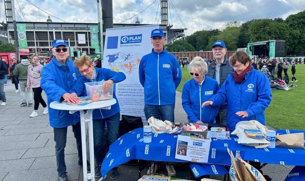 Sechs Personen in blauen Jacken und Hüten mit Plan-Logos stehen hinter einem Tisch mit Informationsmaterial bei einer Veranstaltung im Freien. Im Hintergrund sind ein Stadion und eine Menschenmenge zu sehen.