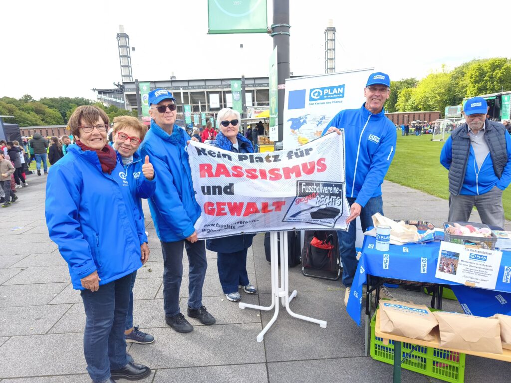 Eine Gruppe von Personen in blauen Jacken steht bei einer Veranstaltung im Freien an einem Tisch und hält ein Transparent mit der Aufschrift Kein Platz für Rassismus und Gewalt. Eine Person zeigt den Daumen nach oben.