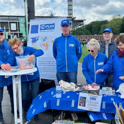Sechs Personen in blauen Jacken und Hüten mit Plan-Logos stehen hinter einem Tisch mit Informationsmaterial bei einer Veranstaltung im Freien. Im Hintergrund sind ein Stadion und eine Menschenmenge zu sehen.