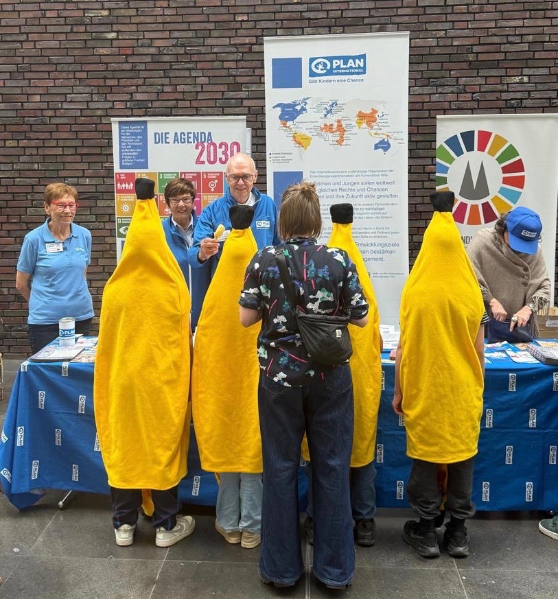Mehrere Personen in Bananenkostümen stehen vor einem Stand, der von Freiwilligen in blauen Hemden betreut wird. Auf dem Tisch und dahinter liegen Informationsplakate und Broschüren aus, darunter auch Materialien von Plan International und der Agenda 2030.