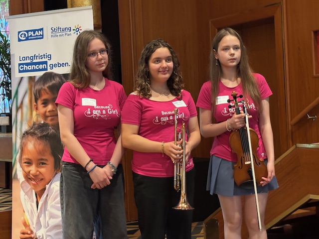Drei Mädchen stehen in einem Raum und tragen jeweils ein einheitliches rosa „Because I am a Girl“-T-Shirt. Eine hält eine Trompete in der Hand, eine andere eine Geige. Auf einem Banner neben ihnen sind Kindergesichter und das Logo von Plan International zu sehen.