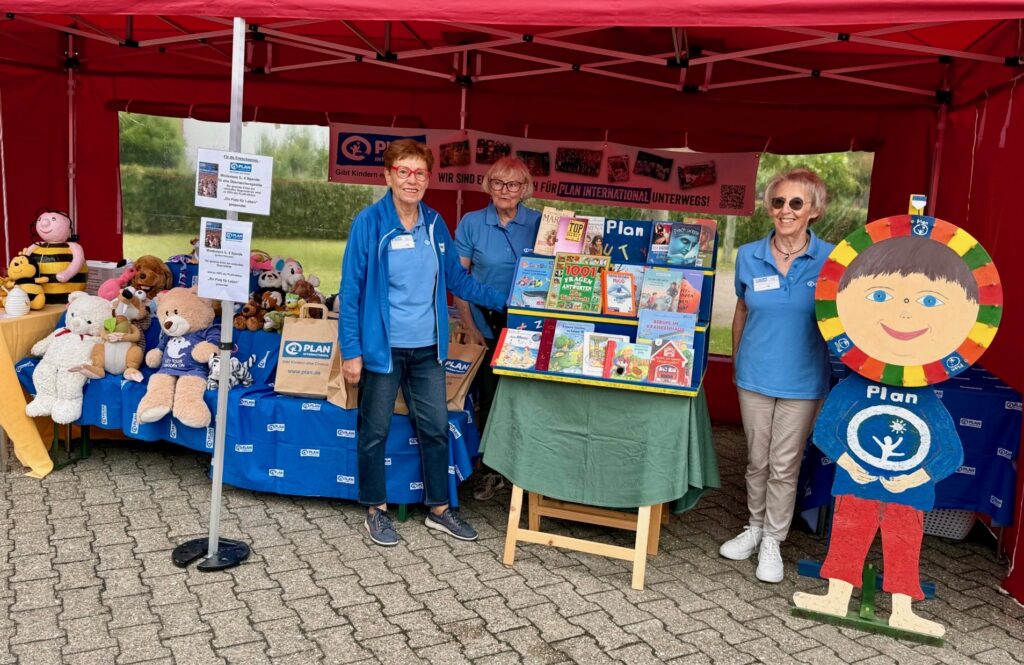 Drei Frauen stehen an einem bunten Wohltätigkeitsstand unter einem roten Zelt mit Tischen, auf denen Stofftiere, Bücher und Informationsmaterial ausliegen. Eine große, fröhliche Kinderpuppe aus Pappe steht am Eingang. Banner und Plakate schmücken den Stand.