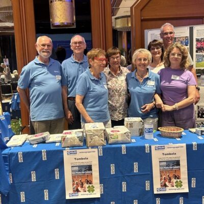 Eine Gruppe von acht lächelnden Senioren, von denen einige blaue Hemden tragen, steht hinter einem mit einem blauen Tuch bedeckten Tisch, auf dem Tombola-Preise und Plakate für eine Tombola-Veranstaltung ausliegen, in einem warm beleuchteten Raum mit Holzvertäfelung und Anschlagtafeln.