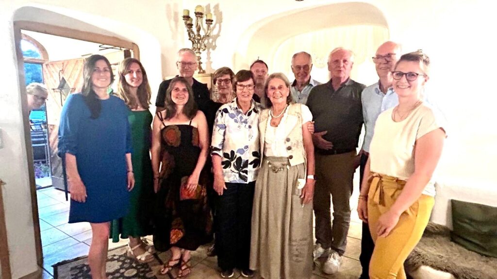 Eine Gruppe von dreizehn Erwachsenen posiert und lächelt gemeinsam in einem warm beleuchteten Raum mit gewölbten Türöffnungen und einem Kerzenleuchter im Hintergrund. Die meisten sind leger gekleidet; einige tragen eine Brille.