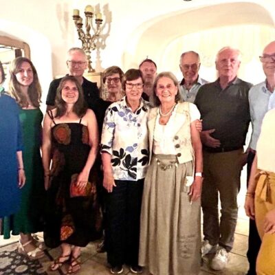 Eine Gruppe von dreizehn Erwachsenen posiert und lächelt gemeinsam in einem warm beleuchteten Raum mit gewölbten Türöffnungen und einem Kerzenleuchter im Hintergrund. Die meisten sind leger gekleidet; einige tragen eine Brille.