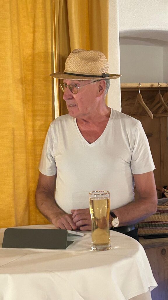 Ein älterer Mann mit einem Strohhut und einem weißen T-Shirt steht an einem runden Tisch, vor sich ein Glas helles Bier. Im Hintergrund sind gelbe Vorhänge und eine Garderobe aus Holz zu sehen.