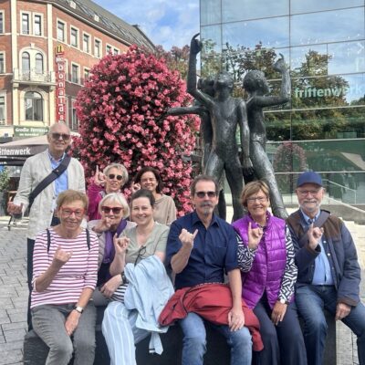 Eine Gruppe von neun Personen sitzt und steht vor einem modernen Glasgebäude, einem rosa blühenden Baum und einer Bronzestatue, lächelt und macht Handzeichen. Es scheint sich um eine lebhafte städtische Kulisse zu handeln.