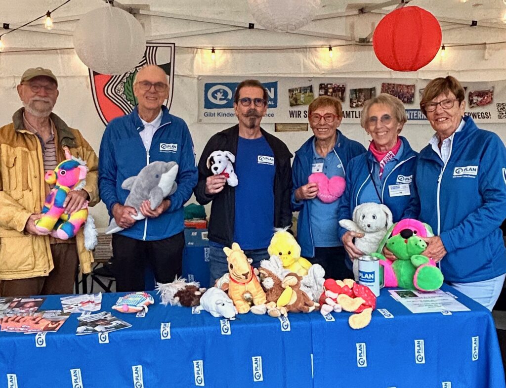 Sechs Erwachsene stehen hinter einem Tisch, der mit bunten Stofftieren und Flyern bedeckt ist, an einem Stand im Freien. Die meisten tragen blaue Jacken und Namensschilder. Der Stand ist mit Papierlaternen und Bannern geschmückt.