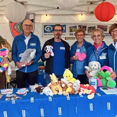 Sechs Erwachsene stehen hinter einem Tisch, der mit bunten Stofftieren und Flyern bedeckt ist, an einem Stand im Freien. Die meisten tragen blaue Jacken und Namensschilder. Der Stand ist mit Papierlaternen und Bannern geschmückt.