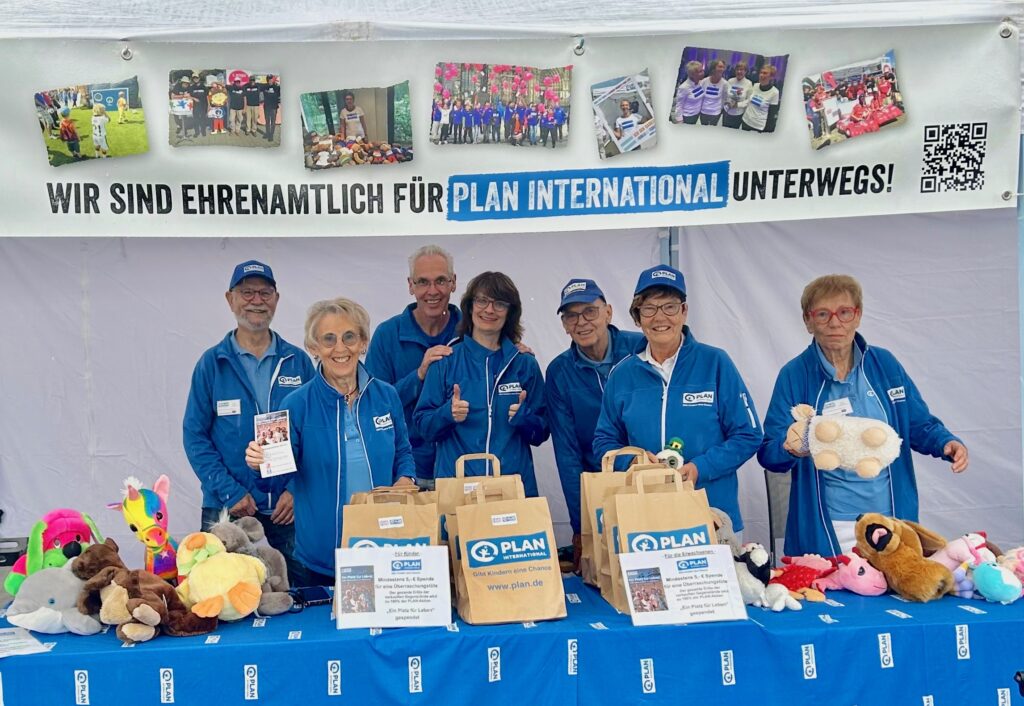 Sieben lächelnde Freiwillige in blauen Jacken stehen an einem Stand mit Spielzeug, Geschenktüten und Broschüren. Auf einem Banner darüber steht: „Wir sind ehrenamtlich für Plan International unterwegs!“.