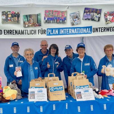 Sieben lächelnde Freiwillige in blauen Jacken stehen an einem Stand mit Spielzeug, Geschenktüten und Broschüren. Auf einem Banner darüber steht: „Wir sind ehrenamtlich für Plan International unterwegs!“.