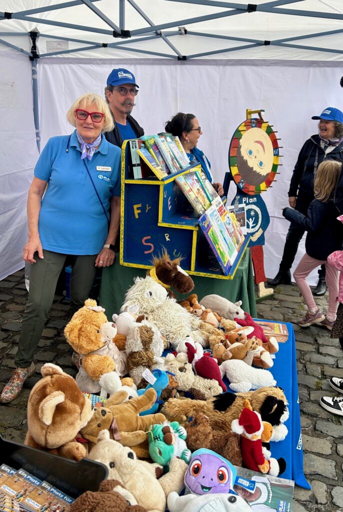 Vor drei Erwachsenen, die hinter einem bunten Spielstand unter einem Zelt bei einer Veranstaltung im Freien stehen, befindet sich ein Tisch mit Stofftieren. In der Nähe sind Kinder zu sehen. Der Boden besteht aus Kopfsteinpflaster.