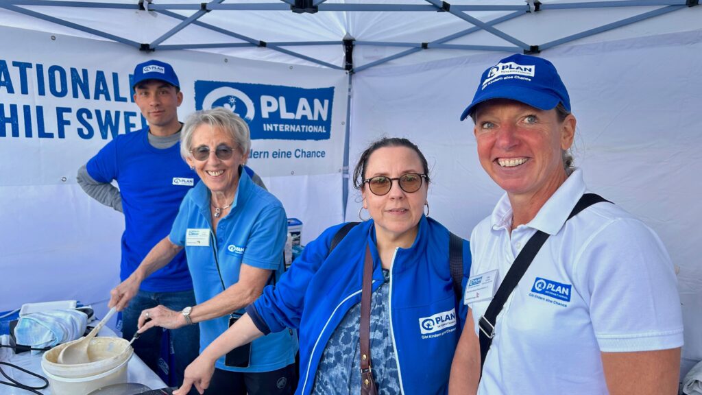 Vier Personen, die blaue T-Shirts und Mützen von Plan International tragen, stehen gemeinsam an einem Stand und lächeln in die Kamera. Hinter ihnen ist ein Banner von Plan International zu sehen. Sie scheinen sich auf einer Veranstaltung oder Messe zu befinden.