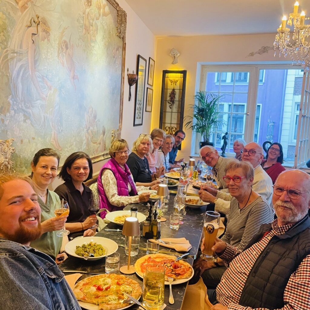 Eine Gruppe von Menschen sitzt in einem gemütlichen, hell beleuchteten Restaurant an einem langen Tisch und genießt gemeinsam Essen und Getränke. Auf dem Tisch stehen Pasta- und Pizzagerichte, und alle lächeln in die Kamera.