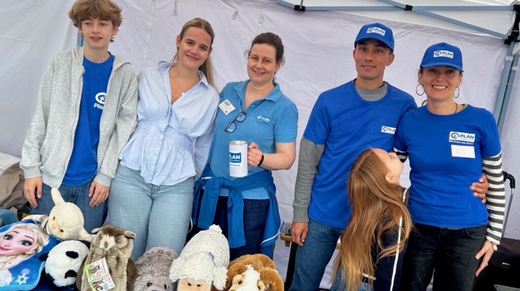 Fünf Erwachsene und ein Kind stehen an einem Stand, auf dessen Tisch Stofftiere liegen. Vier Personen tragen einheitliche blaue T-Shirts und Mützen mit dem PLAN-Logo. Die Gruppe lächelt in einem weißen Zelt.