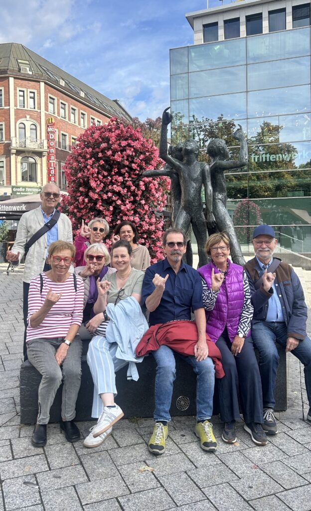 Eine Gruppe von zehn Senioren posiert vor zwei identischen Statuen und einem großen, rosa blühenden Baum auf einem Stadtplatz. Einige strecken ihren kleinen Finger in die Höhe. Im Hintergrund sind Geschäfte und Glasgebäude zu sehen.