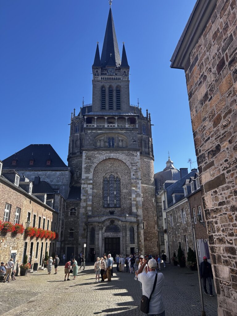 An einem sonnigen Tag stehen Menschen vor einer großen, historischen Steinkirche mit hohem Turm, die von alten Backsteingebäuden umgeben ist, und machen Fotos.