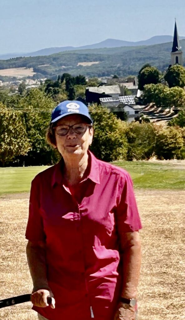 Eine Frau mit einer blauen Baseballkappe und einem leuchtend rosa Shirt steht auf einem grasbewachsenen Golfplatz, im Hintergrund sind ein Dorf und Hügel zu sehen.