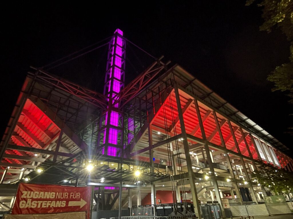 Außenansicht eines modernen Stadions bei Nacht, beleuchtet von leuchtend roten und violetten Lichtern, im Vordergrund ein rotes Transparent mit der Aufschrift ZUGANG NUR FÜR GÄSTEFANS und einem nach rechts zeigenden Pfeil.