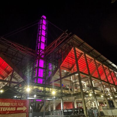 Ein modernes Stadion, das nachts in rotem und violettem Licht erstrahlt, mit einem Schild, auf dem ZUGANG NUR FÜR GÄSTEFANS steht, versehen mit einem Pfeil.