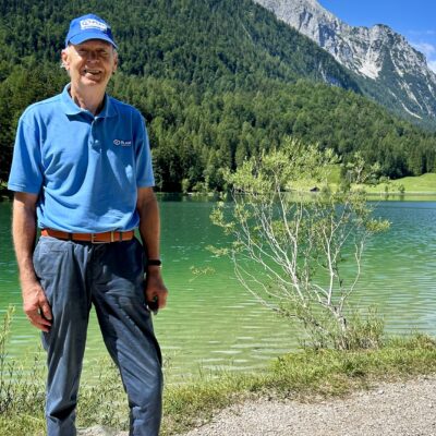 Ein Mann in einem blauen Poloshirt und einer Mütze steht auf einem Schotterweg neben einem türkisfarbenen See, im Hintergrund sind bewaldete Berge zu sehen.