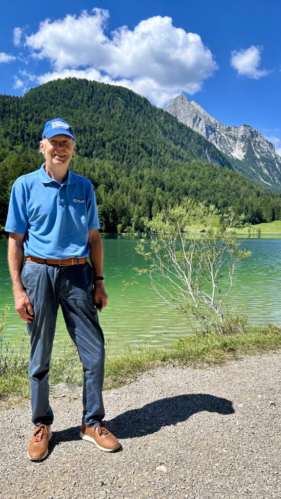 Ein Mann in einem blauen Poloshirt und einer Mütze steht auf einem Schotterweg neben einem türkisfarbenen See, im Hintergrund sind bewaldete Berge zu sehen.