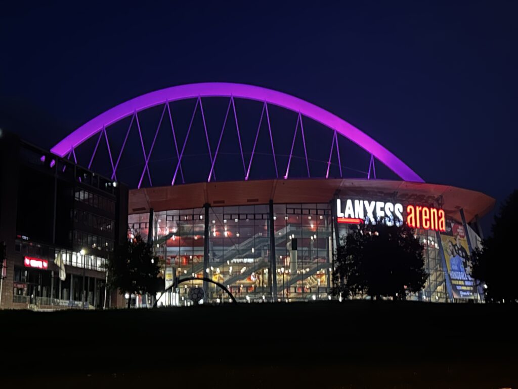 Die Lanxess Arena bei Nacht, mit einem großen, hell erleuchteten violetten Bogen über dem Dach.
