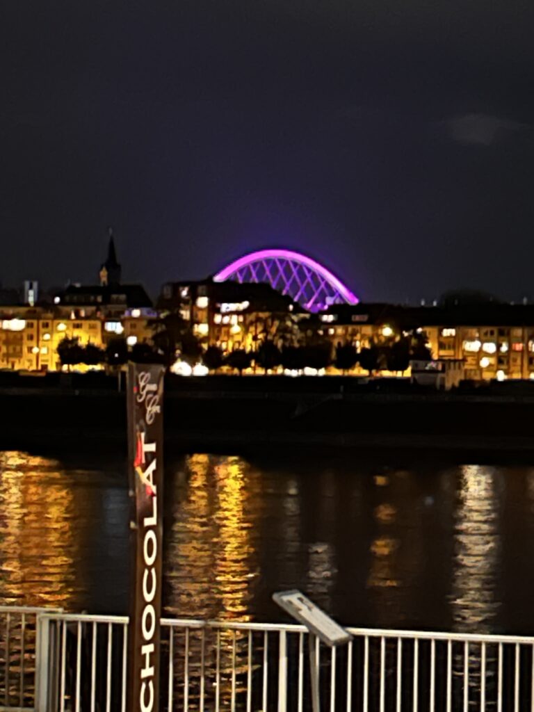 Eine nächtliche Skyline mit einer leuchtend violetten Bogenbrücke, die sich in einem dunklen Fluss spiegelt.